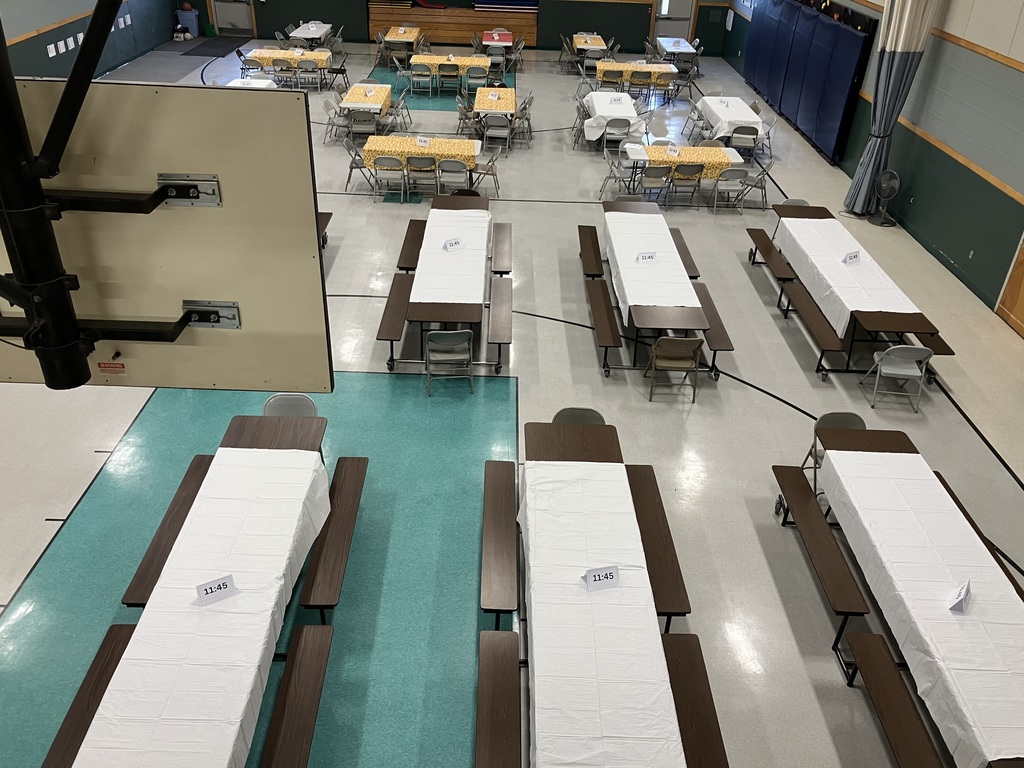 A high-angle view looking across the gymnasium, showcasing the extensive table setup for the meal. The tables are arranged in sections, with some covered in white cloths and others in yellow. A basketball hoop is visible at the far end of the gym, and a large black structural support column and beige partition dominate the right side of the foreground.