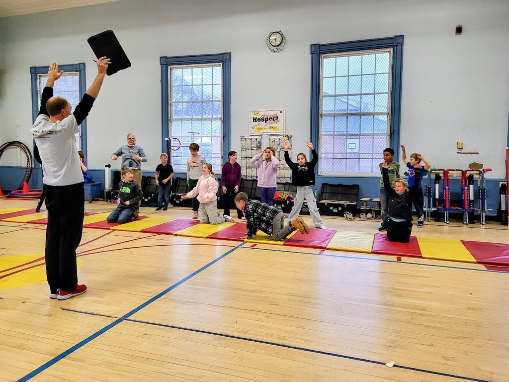 A PE instructor stands in front of a group of elementary students in a gym, holding a black foam block above his head. The students kneel or stand on colorful mats, raising their hands and reacting enthusiastically while participating in an activity.