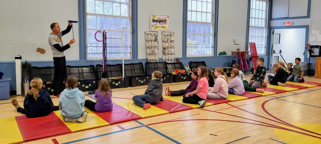 A PE instructor demonstrates how to spin a plate on a stick while a group of elementary students sit on red and yellow mats watching attentively. Gym equipment and large windows line the background.