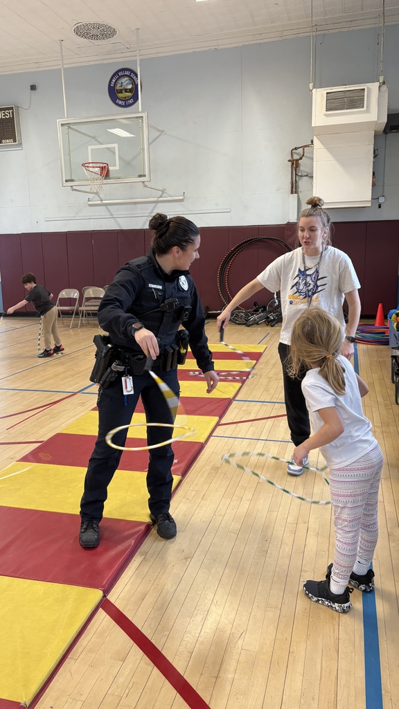 A police officer in uniform and a PE teacher help a young student use a spinning rope toy in the gym. Other students play with similar equipment in the background.