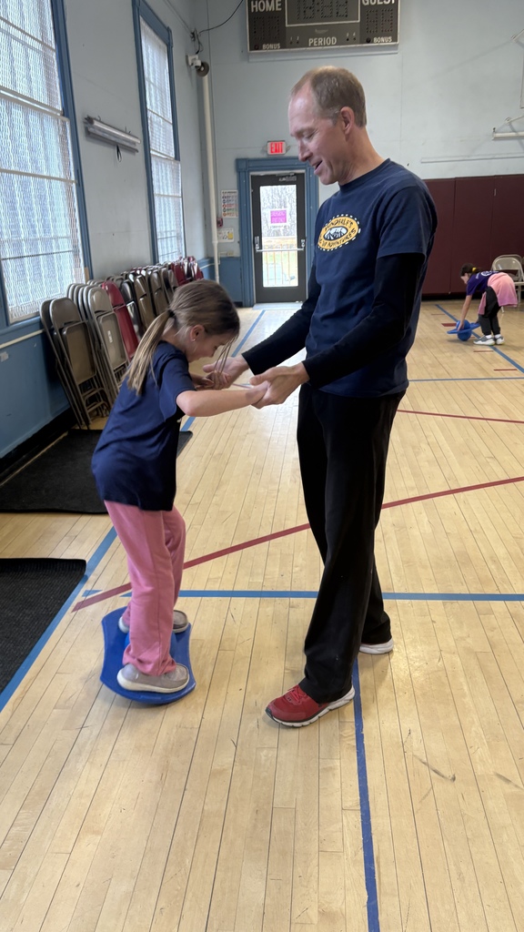 A PE instructor helps a young girl balance on a wobble board in the gym, holding her hands for support. She smiles while trying to stabilize herself.