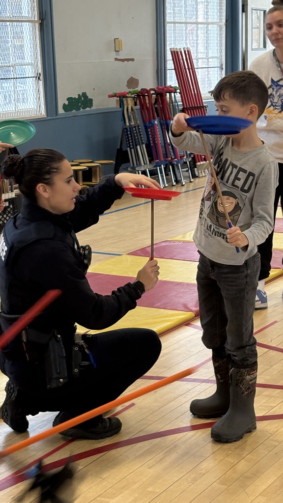 A police officer kneels on a gym mat, helping a young student balance a spinning plate on a stick. The student holds a blue plate while the officer demonstrates with a red one. Gym equipment is stacked behind them.