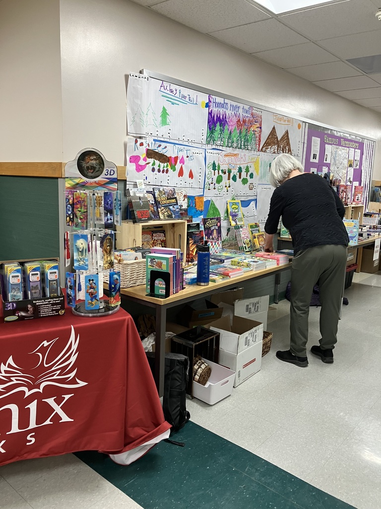 A display table for the book fair is set up beneath a bulletin board titled "Famous Vermonters" which features a map of Vermont. The table is filled with various books arranged on metal tiered racks and laid flat, with a sign indicating the section is for "MIDDLE READERS".