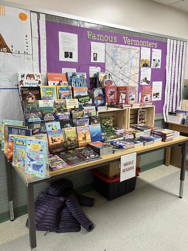 IMG_2904.jpg: Four adult women, presumably staff or volunteers, are standing behind a table covered with a red Phoenix Books tablecloth and holding up children's books. They are smiling at the camera, promoting the book fair. A wooden crate sits on the table, which also displays small gift items and a sign indicating payment methods. IMG_2895 (1).jpg: A close-up of the Phoenix Books book fair display table. The red tablecloth features the store's logo. A wooden crate on top holds a sign detailing accepted payment methods (Visa, MC, Checks & Cash). A rotating display rack features 3D novelties and gifts. IMG_2897.jpg: A display table for the book fair is set up beneath a bulletin board titled "Famous Vermonters" which features a map of Vermont. The table is filled with various books arranged on metal tiered racks and laid flat, with a sign indicating the section is for "MIDDLE READERS". IMG_2899.jpg: A long wooden table set up in a school hallway with books displayed for a book fair. A tiered metal rack holds numerous titles, and the table displays stacks of books underneath a sign labeled "EARLY READERS". A red fire extinguisher is visible on the wall in the background. IMG_2898.jpg: A table displaying books for the book fair, categorized as "EARLY READERS". The books are laid out on the table and arranged in white baskets and small wooden shelving units. A purple chart showing time measurements is visible on the wall to the left. IMG_2900.jpg: A close-up shot of the cover of a non-fiction children's book titled "DIRT BIKES" in large, red and white letters. The cover features an image of a person riding a dirt bike in racing gear. IMG_2901.jpg: A close-up shot of two book covers: the top book is a question-based title asking, "BUT WHY ARE LLAMAS TICKLISH?" The bottom book cover is a sports title titled "SUPERCROSS BUMPS AND JUMPS." Classroom Photos IMG_2861 (1).jpg: A boy wearing glasses is standing next to a black pocket chart calendar, with his arm raised in a possible wave or question. The chart displays the days of the week and two small analog clocks labeled '3' and '4'. A girl's arm and a red marker are visible in the foreground. IMG_2863 (1).jpg: A close-up of a student's arm and hand writing on a large sheet of paper titled "Calendar Grid Observations." The chart has columns for Date, Time, am/pm, Between today + yesterday, and Total time elapsed since 11/1, used for a lesson on calculating elapsed time.