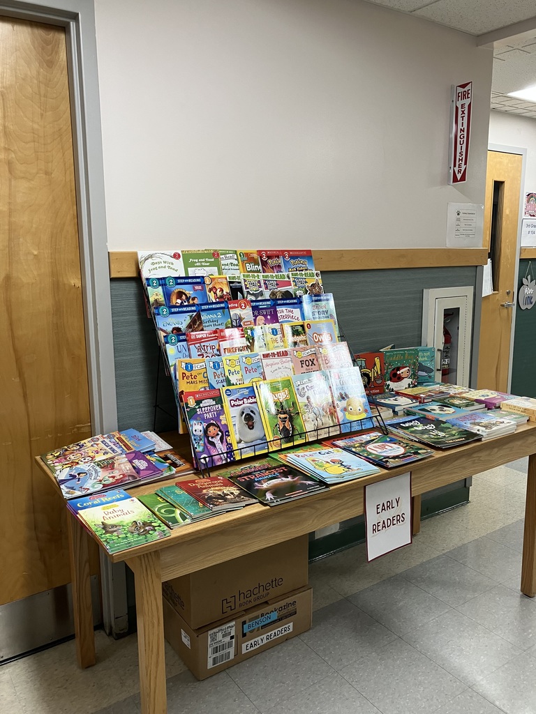 A long wooden table set up in a school hallway with books displayed for a book fair. A tiered metal rack holds numerous titles, and the table displays stacks of books underneath a sign labeled "EARLY READERS". A red fire extinguisher is visible on the wall in the background.