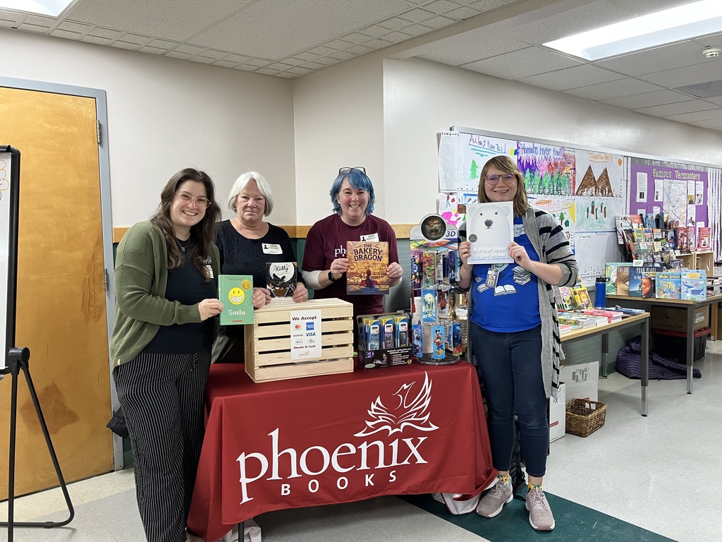 Four adult women, presumably staff or volunteers, are standing behind a table covered with a red Phoenix Books tablecloth and holding up children's books. They are smiling at the camera, promoting the book fair. A wooden crate sits on the table, which also displays small gift items and a sign indicating payment methods.