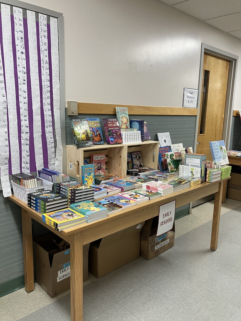 A table displaying books for the book fair, categorized as "EARLY READERS". The books are laid out on the table and arranged in white baskets and small wooden shelving units. A purple chart showing time measurements is visible on the wall to the left.