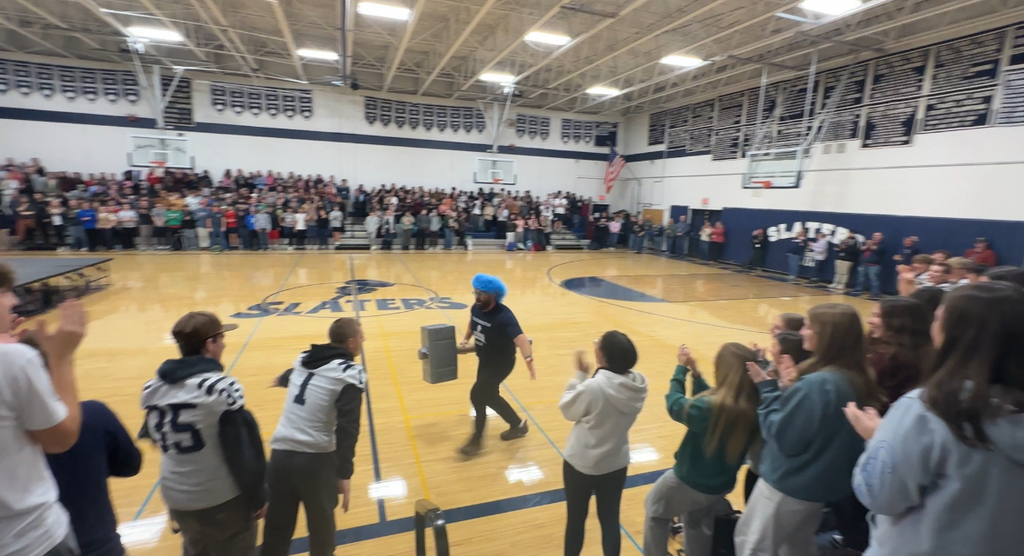 A staff member wearing a bright blue wig runs playfully across the gym floor holding a large prop hammer as students and football players cheer and clap around him. The bleachers are filled with students standing and watching the pep rally.