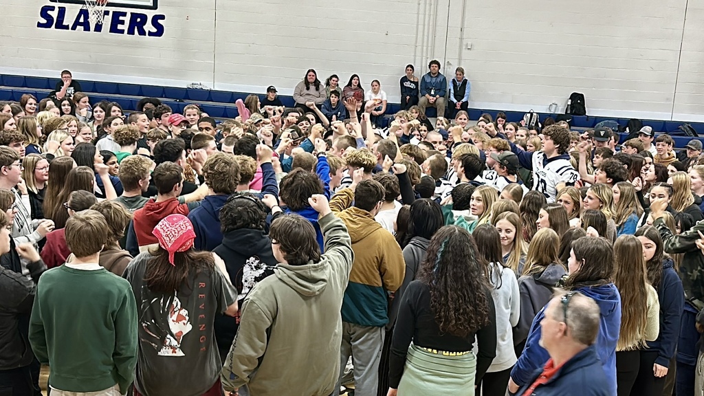 A large group of high school students gathers tightly in a circle on the gym floor with their fists raised in the air, cheering together. Several football players in Slaters jerseys are visible in the crowd, and additional students sit on the bleachers behind them.
