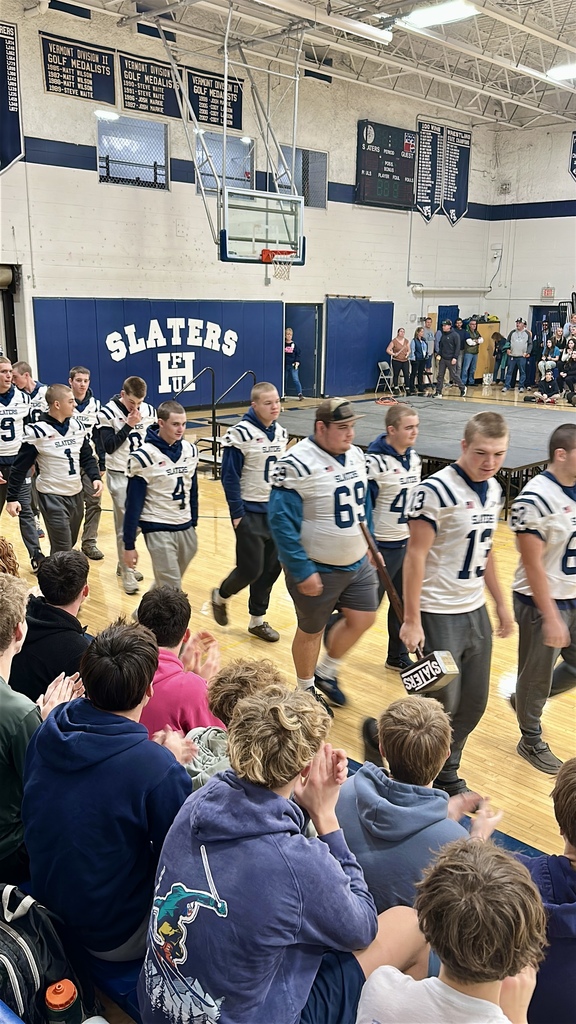 High school football players wearing white Slaters jerseys walk in a line through the gym during a pep rally as students seated on the bleachers clap and cheer. Banners, a basketball hoop, and the Slaters logo are visible in the background.