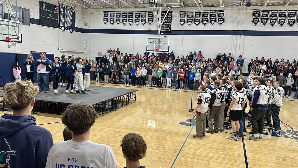 Football players in Slaters jerseys stand together facing a raised stage while a large crowd of students fills the bleachers behind them. Staff members on stage cheer and clap during the pep rally in the school gym, decorated with championship banners.