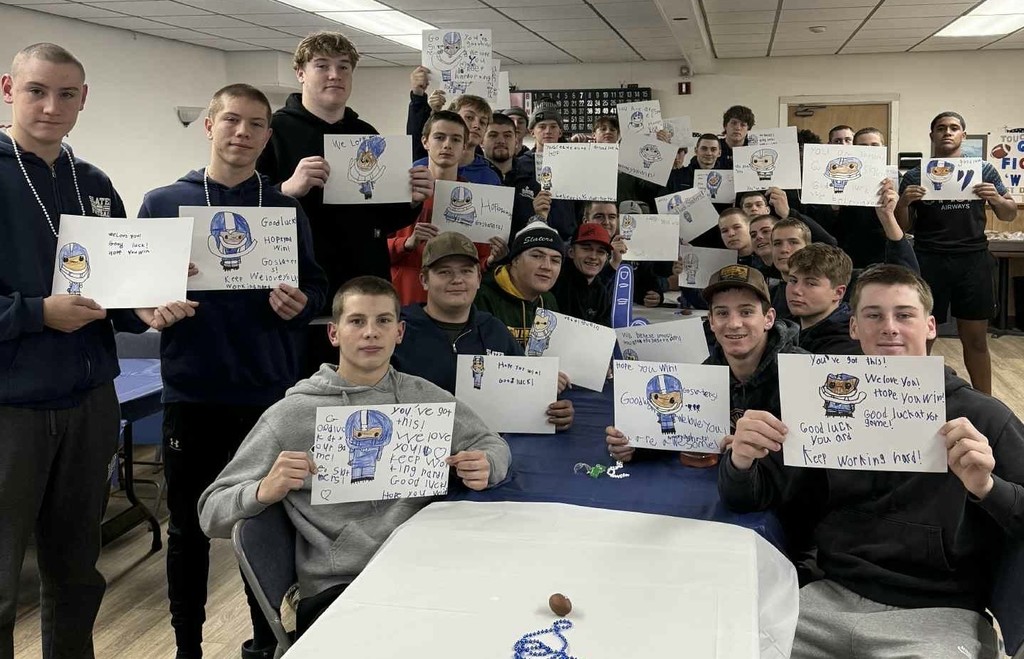 A large group of high school football players sit and stand together in a team room, each holding up a handmade good-luck poster created by younger students. The posters feature drawings of a Slaters football player and encouraging messages like “Good luck!”, “You’ve got this!”, and “Go Slaters!” The players smile proudly as they display the artwork on the tables and in their hands.