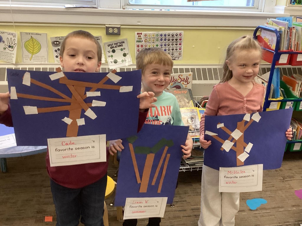 Three young students stand in a classroom holding up artwork made from cut paper. Each picture shows a tree made of brown paper strips with white paper pieces representing snow. Below each artwork is a sentence starter indicating the child’s favorite season. The students smile as they display their winter-themed trees.