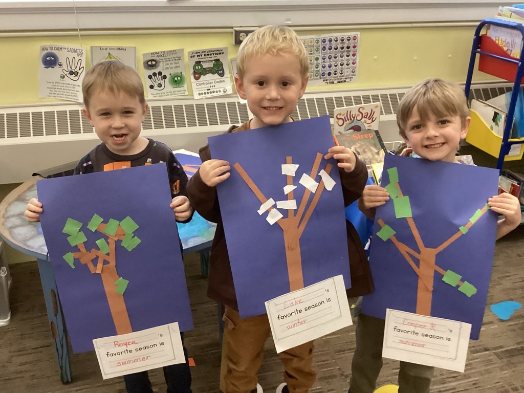 Three young students stand in a classroom holding up paper-collage tree artwork. Two of the trees have green paper leaves representing summer, while one has white paper pieces representing winter. Each artwork includes a label stating the child’s favorite season. The students smile proudly at the camera.