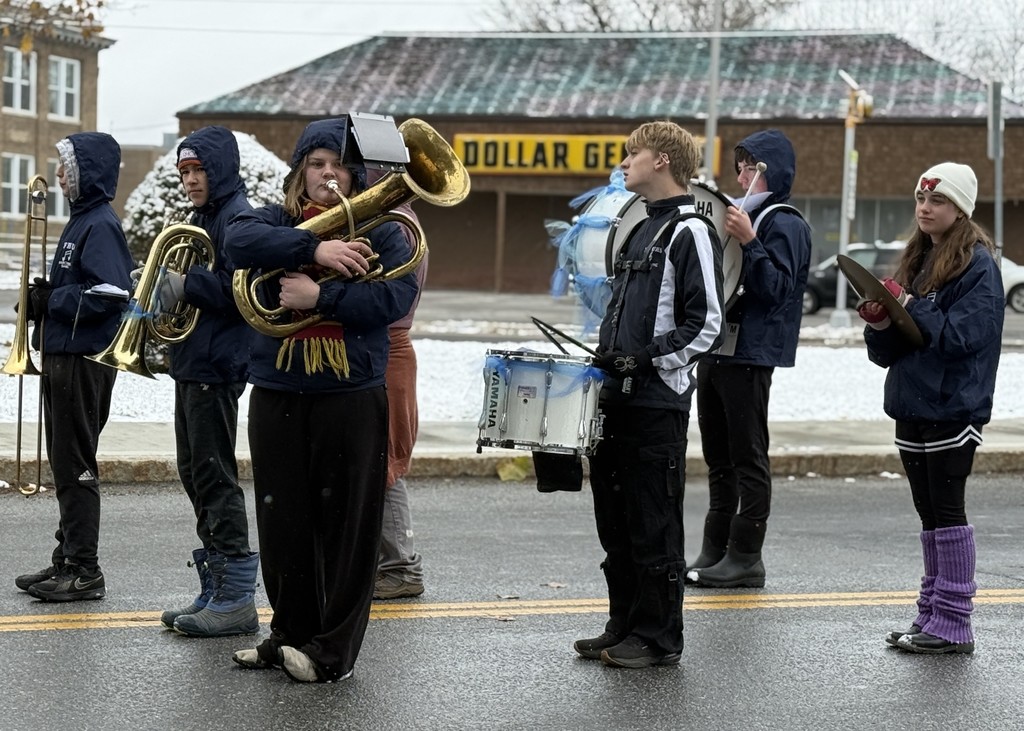 FHU marching band percussion and brass players stand in formation during the parade on a snowy day.