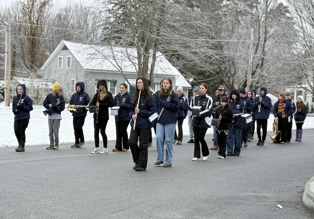 FHU marching band students march in formation along a snowy residential street while playing instruments.