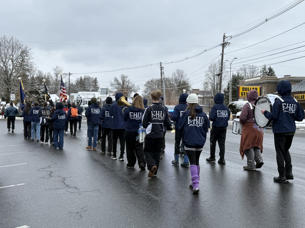 Students in FHU marching band uniforms walk down a street in a parade on a cold, overcast day, carrying instruments and following the color guard.