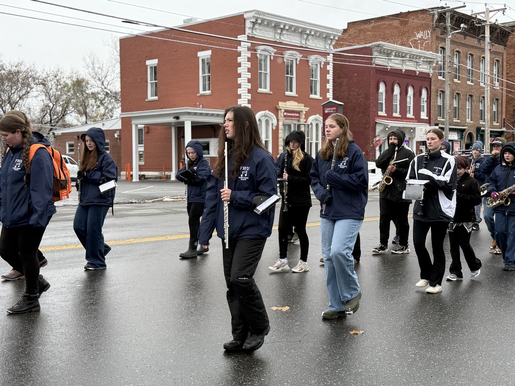 FHU marching band students walk in formation through town, playing instruments while passing brick buildings.