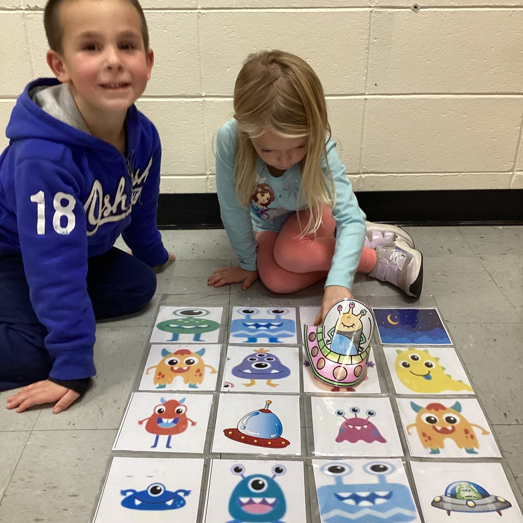 Two children sit on the floor with a grid of monster pictures, one child smiling toward the camera while the other moves a small paper alien spaceship on the cards.