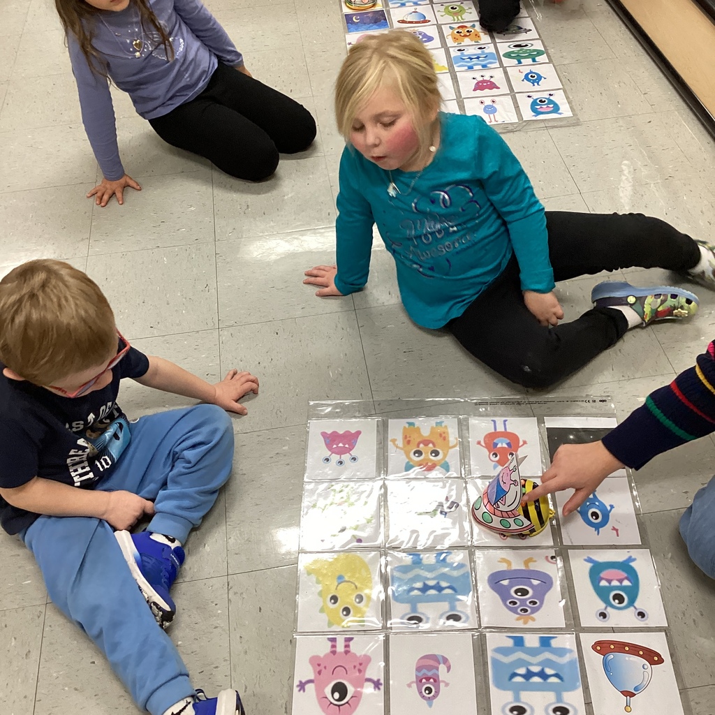 Two young children sit on the floor looking at a grid of colorful monster cards while an adult hand points to a small paper alien spaceship on the grid.