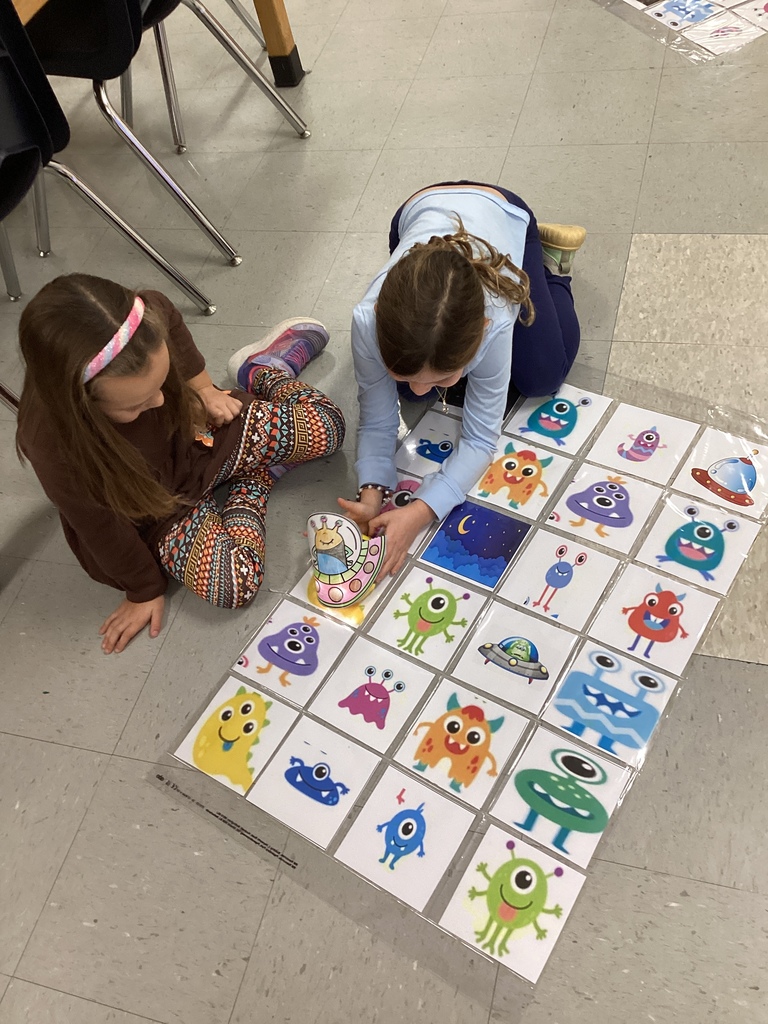 Two girls sit on the floor working together on a grid of colorful monster images, moving a small paper alien spaceship across the cards.