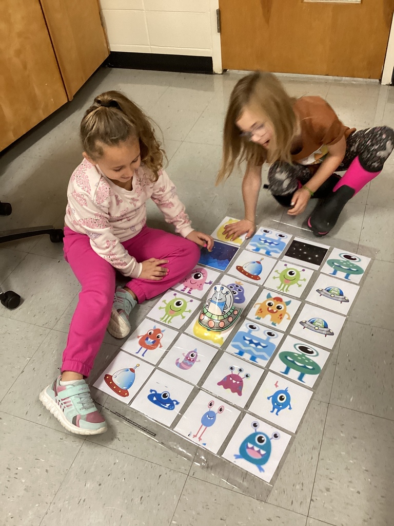 Two young girls sit on the floor laughing and playing with a grid of monster cards, placing a paper alien spaceship on one of the images.