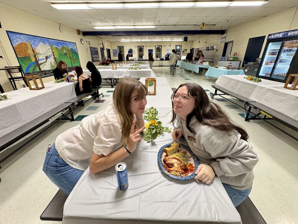 Two students sit at a decorated cafeteria table, posing and smiling while eating spaghetti. One student leans in and playfully bites a spaghetti noodle.