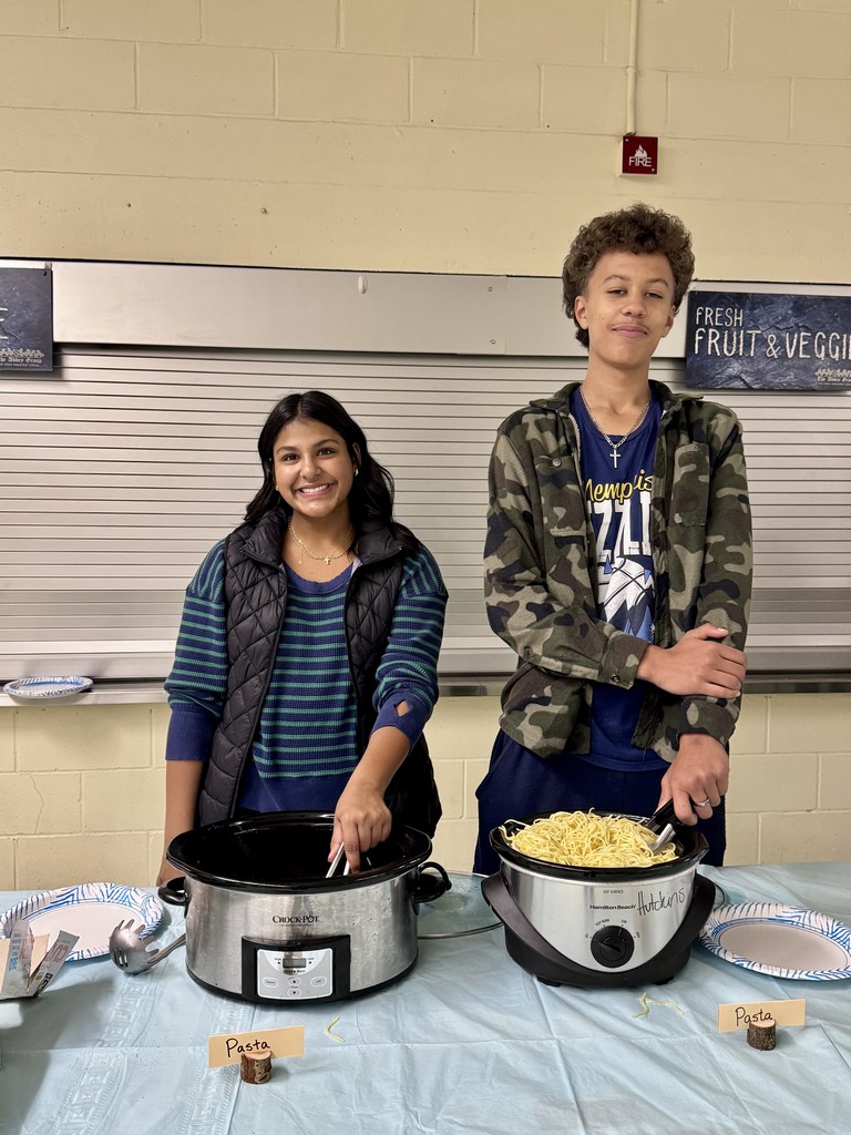 Two students stand behind buffet dishes labeled “Pasta,” smiling as they help serve the dinner. They are positioned in front of a cafeteria serving station.