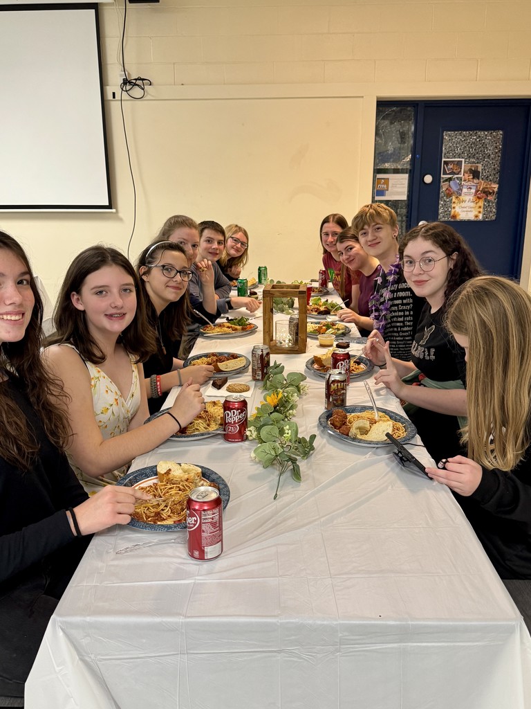 A large group of middle school students sit together at a long table enjoying their spaghetti dinner. They smile at the camera, and the table is decorated with greenery and flowers.