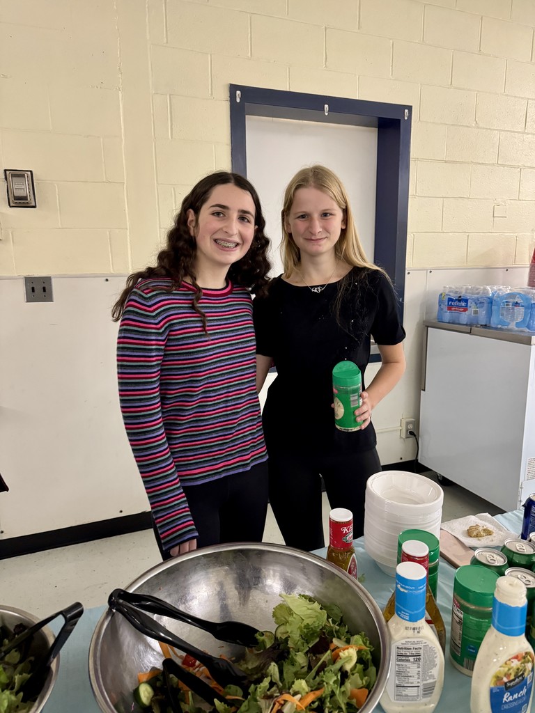 Two students stand behind a large salad bowl at the dinner event, smiling while holding salad dressing containers. A row of dressings and bowls is arranged on the table in front of them.