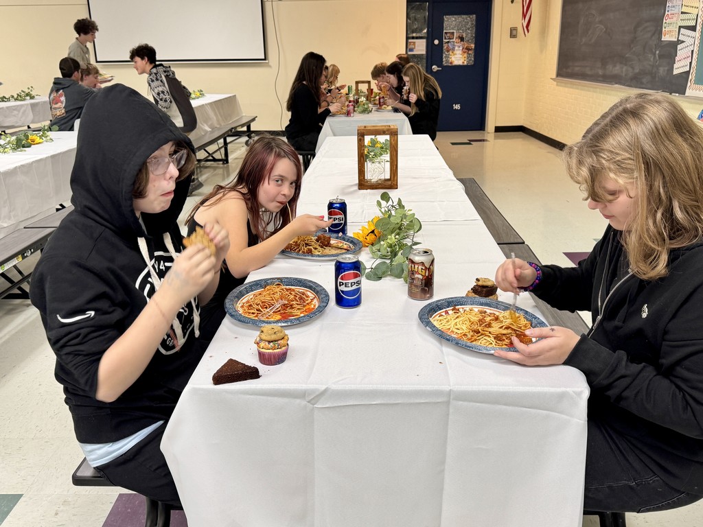 Students sit at a long decorated table eating spaghetti, cupcakes, and brownies during the dinner event. Other students can be seen eating at tables in the background.
