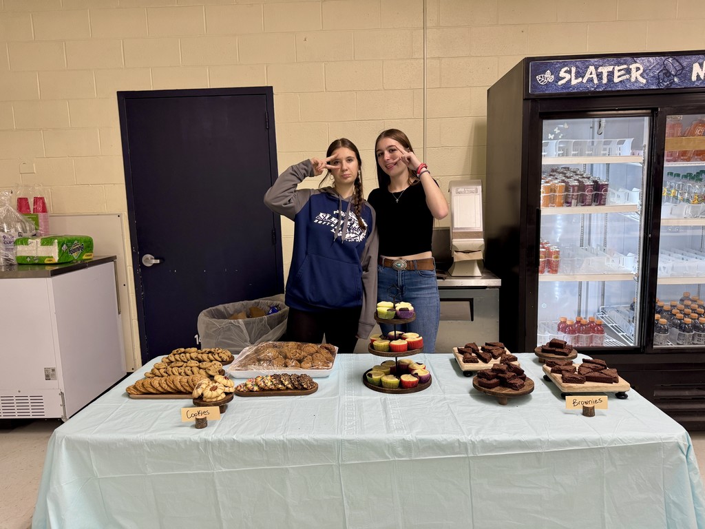 Two middle school students stand behind a dessert table covered with cookies, cupcakes, and brownies. They are posing playfully with peace signs. A refrigerated case labeled “Slater Nation” is visible behind them.