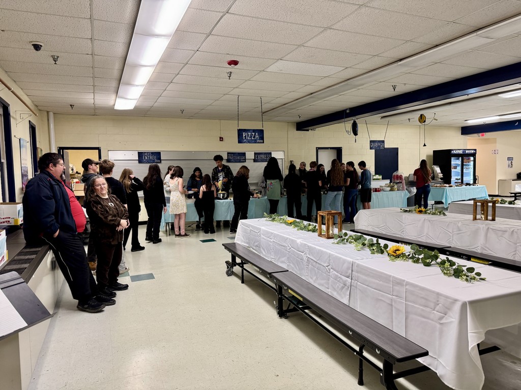 A group of students and a few adults gather around buffet tables in a cafeteria as the dinner event begins. Decorated tables with flowers and lanterns fill the room.