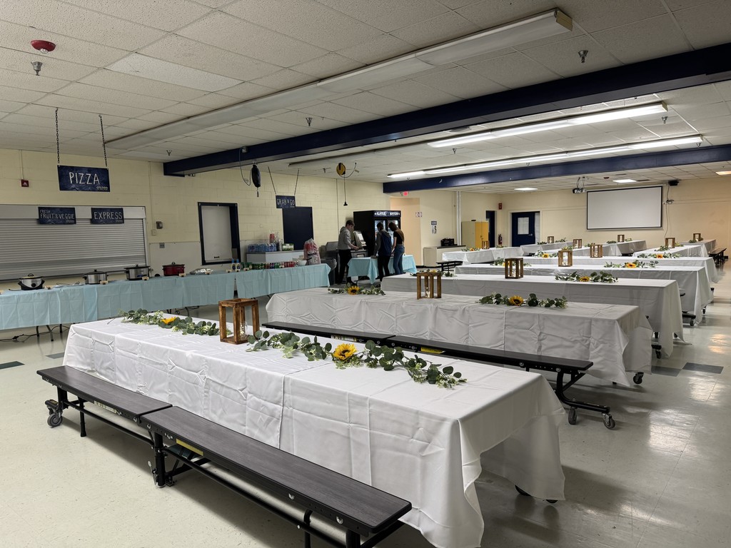 A cafeteria decorated for a dinner event with long tables covered in white tablecloths, floral centerpieces, and wooden lanterns. Buffet tables line the back wall, and a few students and adults are preparing food.