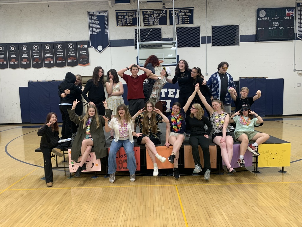 The same cast of high school theater students pose again in the gymnasium, but this time they are laughing, cheering, hugging, and striking playful poses. Students on the front row throw peace signs, while those on the risers celebrate with raised arms and dramatic expressions. The scene is energetic and joyful, showing the cast’s personality and camaraderie.