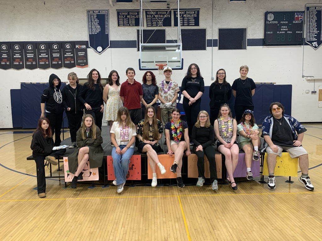 A large group of high school theater students pose on risers in a gymnasium for a cast photo. Some students are dressed in dark clothing, others in Hawaiian shirts, dresses, or casual wear, reflecting their characters from the play Murder Ahoy. They stand and sit in two rows, smiling calmly at the camera. Colorful handmade signs labeled “Class of ’25” are attached to the risers. Banners and a scoreboard hang on the gym wall behind them.