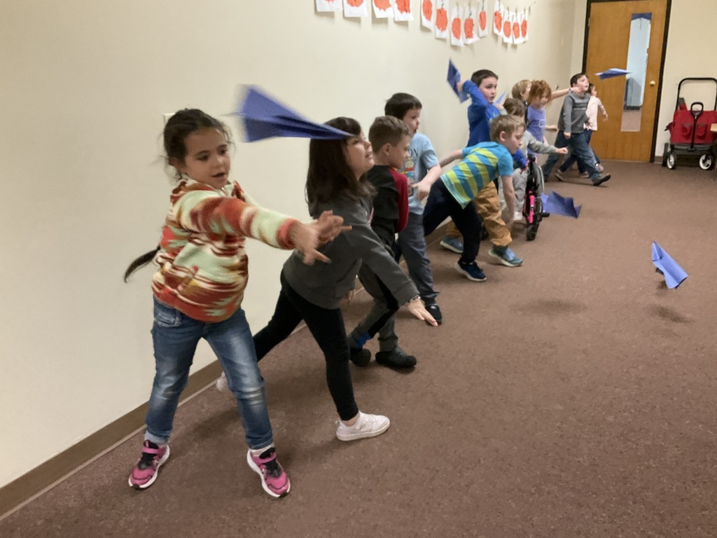 Children in a hallway all throw blue paper airplanes at the same time.