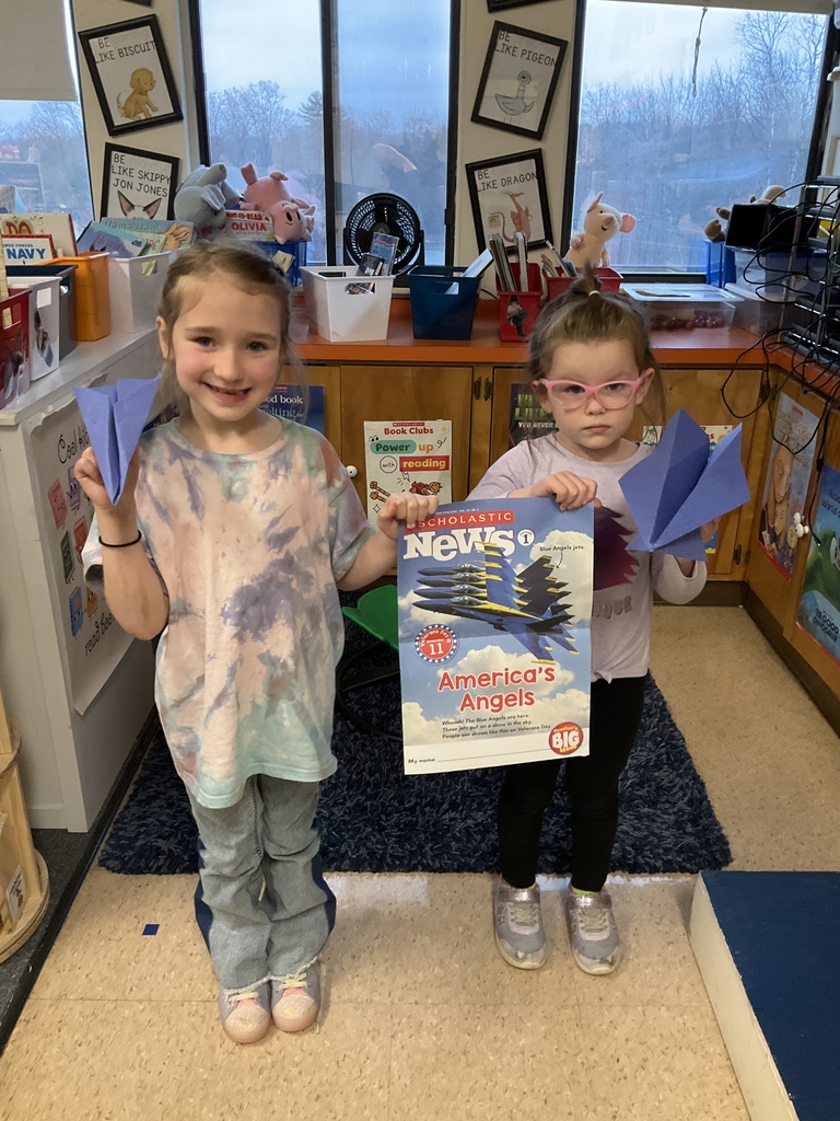 Two young girls stand in a classroom holding blue paper airplanes; one also holds a Scholastic News magazine featuring the Blue Angels.