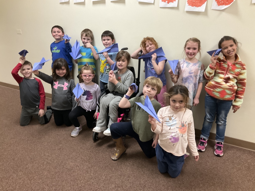 A group of elementary students pose together in a hallway, each holding a blue paper airplane.