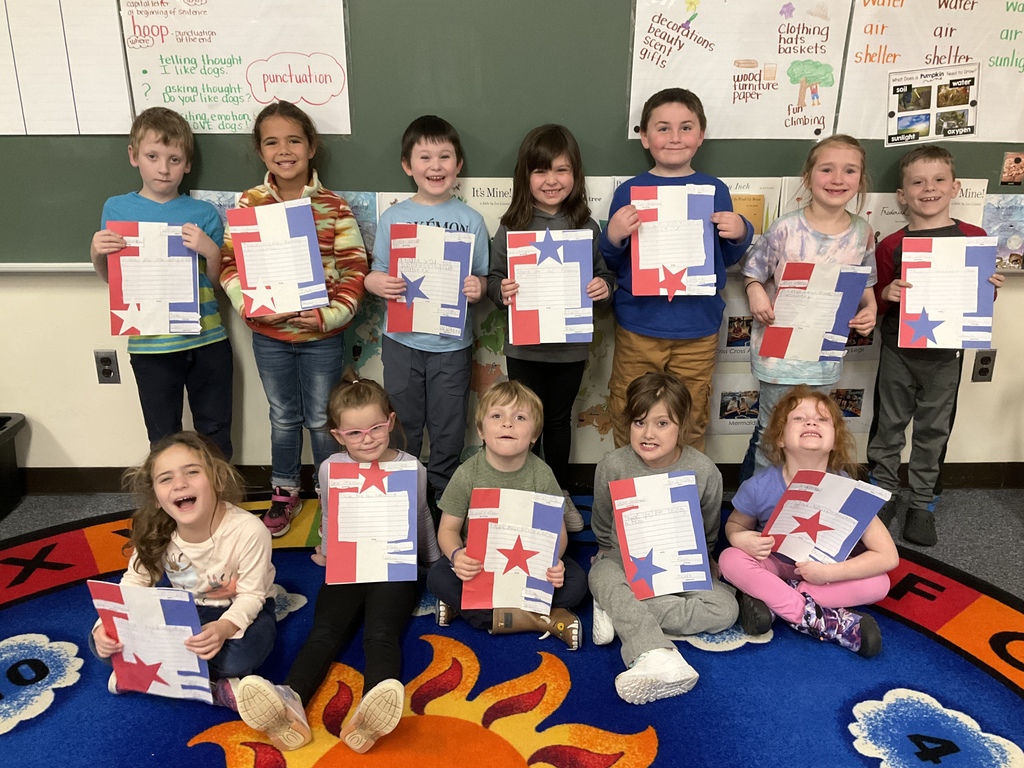 A class of students sits and stands in front of a chalkboard, each holding a patriotic red, white, and blue letter-writing project.