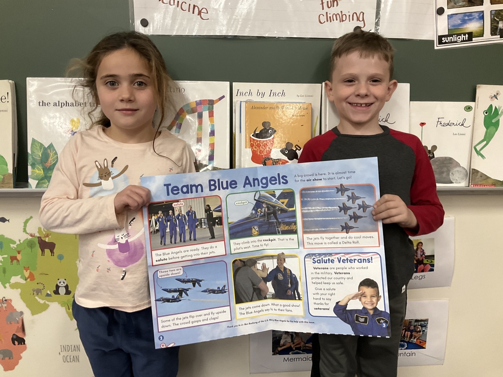 A girl and a boy stand in front of a chalkboard holding open a Scholastic News spread titled “Team Blue Angels.”