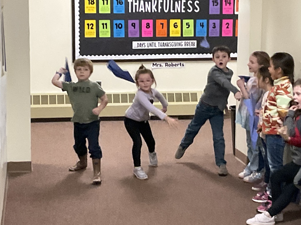 Three children in a hallway throw blue paper airplanes while classmates watch from the side.