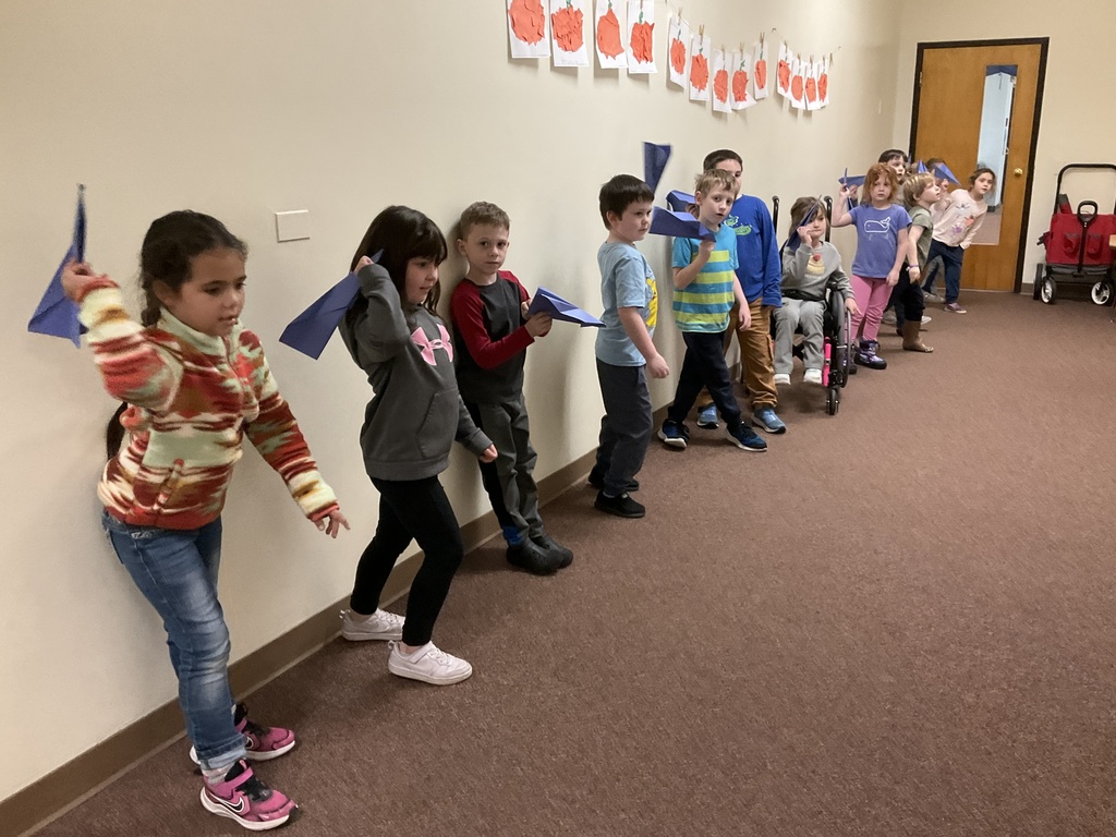 A line of children stand in a hallway holding blue paper airplanes, preparing to throw them.