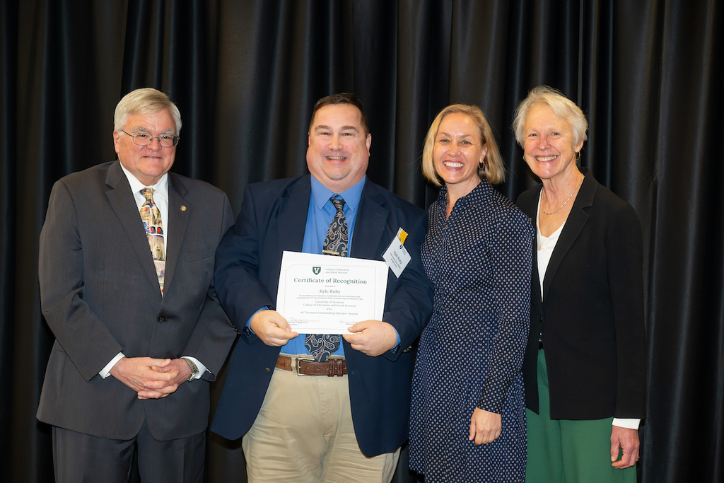Kyle Ruby stands onstage holding his Certificate of Recognition, posed with three award presenters in formal attire in front of a black curtain.