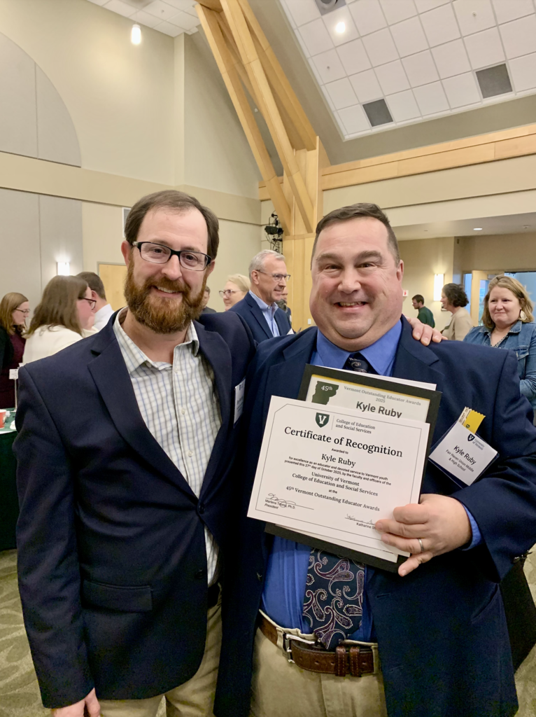 Kyle Ruby smiles while holding his Certificate of Recognition, standing beside a colleague at an indoor awards reception with other attendees in the background.