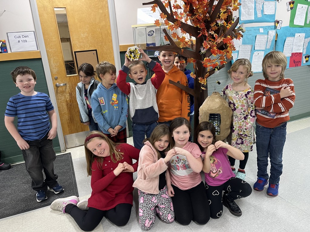 A group of elementary school children pose together in a hallway beside a tall autumn-themed paper tree with colorful leaves and a large paper beehive attached to it. Some children stand behind the tree while others sit or kneel in front, many of them making heart shapes with their hands. One child in the back holds a small craft decorated like a beehive above their head. Student work is displayed on the wall behind them, adding bright colors to the scene.