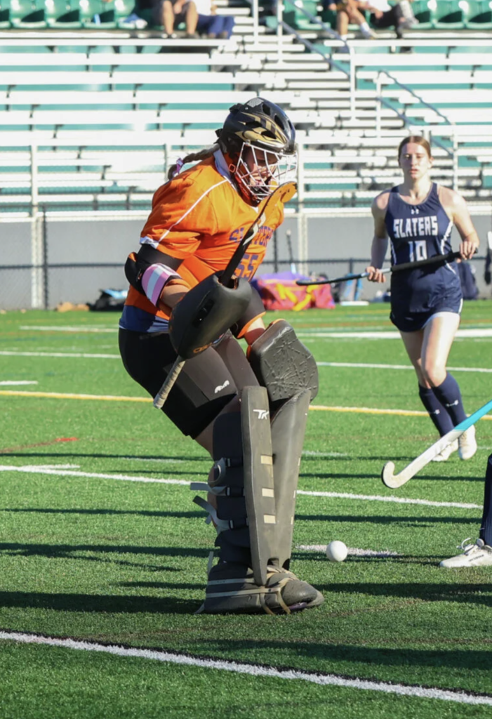 A field hockey goalkeeper in full protective gear, wearing an orange jersey and black pads, steps forward to block the ball during a game. A Slaters player in a navy uniform approaches from behind, while the action takes place on a bright green turf field with empty bleachers in the background.