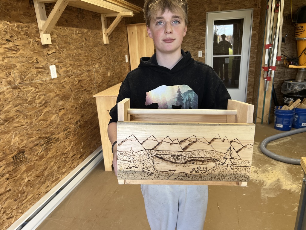 A middle school student stands in a woodshop holding a wooden toolbox he built, decorated with a hand-burned landscape scene of mountains, trees, and a lake. Shelves, wood panels, and shop tools are visible in the background.