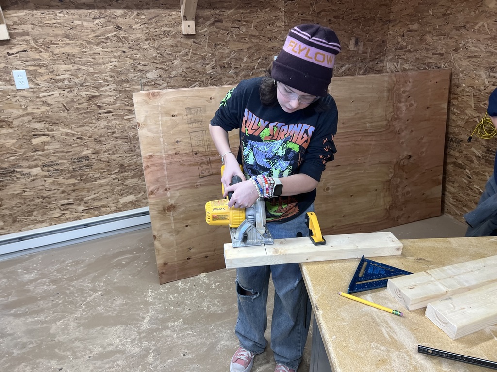 A middle school student wearing a winter hat and graphic T-shirt uses a circular saw to cut a piece of wood in a workshop. Measuring tools, lumber, and plywood sheets are on the table and walls around them.