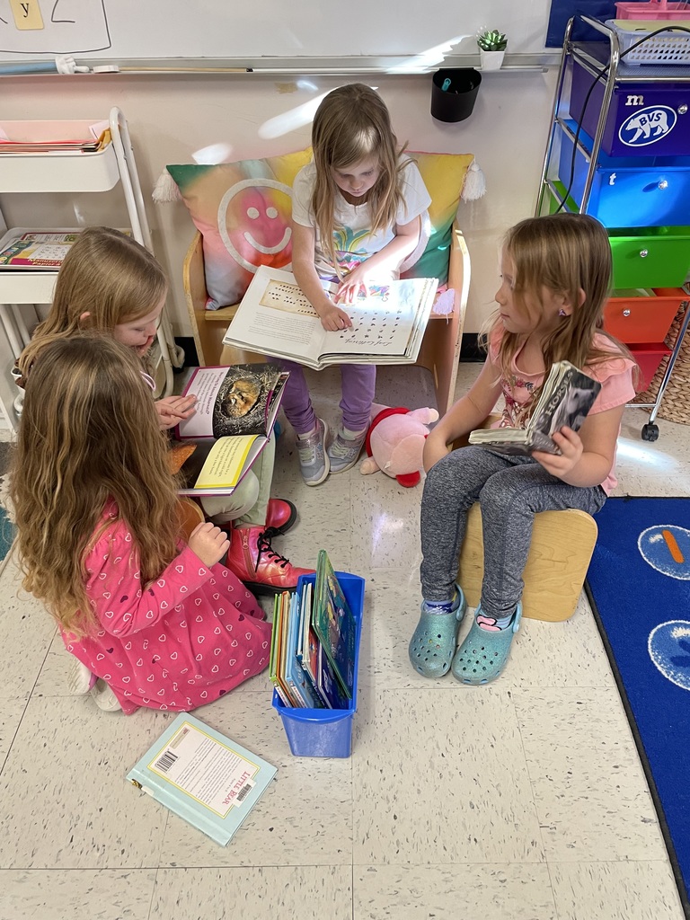 Four young girls gathered in a cozy classroom area. One girl is sitting on a chair/bench with a smiling sun pillow, reading aloud from a large book to the other three who are sitting on the floor, looking at books, suggesting a small reading group or story time. 